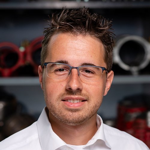 Ein junger Mann mit Brille lächelt freundlich vor einem Hintergrund aus technischen Geräten und Werkzeugen.