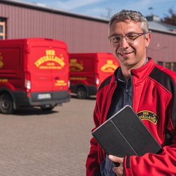Ein lächelnder Mann in einem roten Jackett steht vor mehreren roten Lieferwagen auf einem gepflasterten Hof.