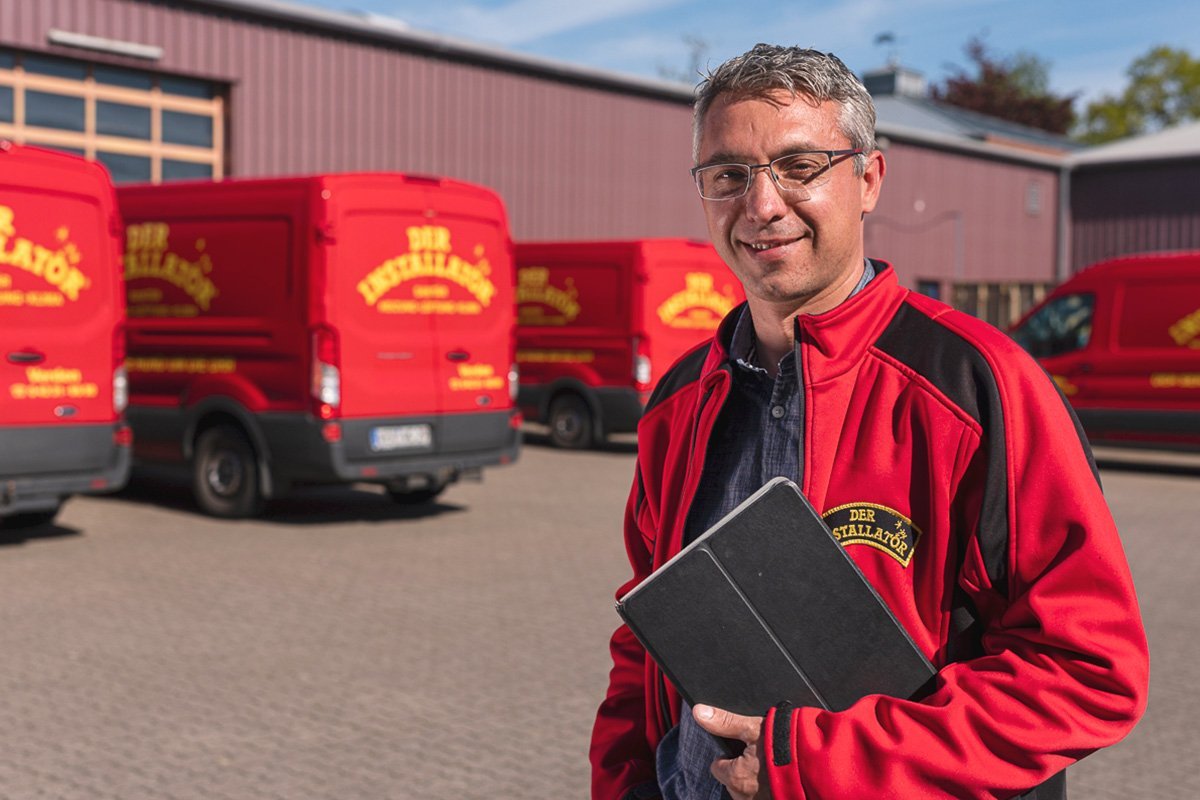 Ein lächelnder Mann in einem roten Jackett steht vor mehreren roten Lieferwagen auf einem gepflasterten Hof.