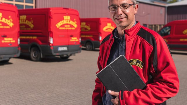 Ein lächelnder Mann in einem roten Jackett steht vor mehreren roten Lieferwagen auf einem gepflasterten Hof.
