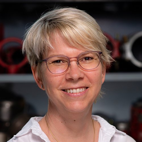 Eine lächelnde Frau mit blonden, kurzen Haaren und Brille steht vor einer Werkstatt mit technischen Geräten im Hintergrund.