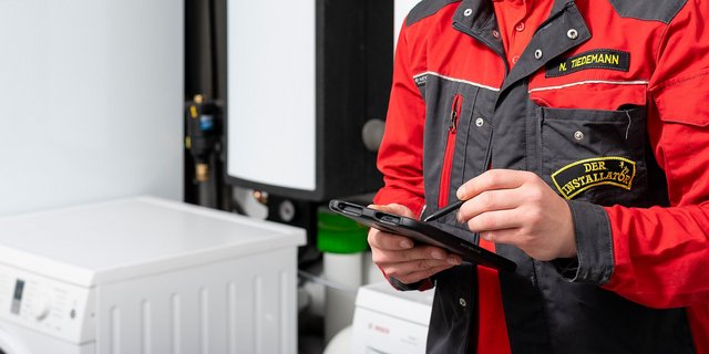 Ein Installateur in einem roten und schwarzen Arbeitsanzug nutzt ein Tablet, während er in einem modernen Technikraum steht.
