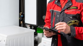 Ein Installateur in einem roten und schwarzen Arbeitsanzug nutzt ein Tablet, während er in einem modernen Technikraum steht.