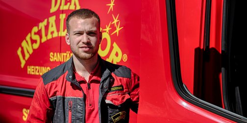 Ein junger Handwerker steht lächelnd vor einem roten Fahrzeug mit dem Schriftzug 'Der Installateur' im Hintergrund.