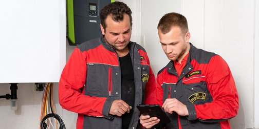 Zwei Handwerker in roten und grauen Arbeitsjacken betrachten gemeinsam ein Tablet in einem hellen Raum mit technischen Geräten.
