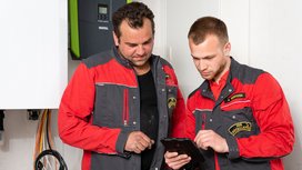 Zwei Handwerker in roten und grauen Arbeitsjacken betrachten gemeinsam ein Tablet in einem hellen Raum mit technischen Geräten.