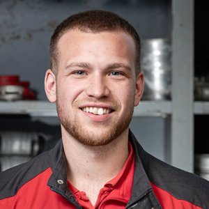 Ein junger Mann mit einem breiten Lächeln trägt eine rote und schwarze Arbeitsjacke vor einem Regal mit Werkzeugen.