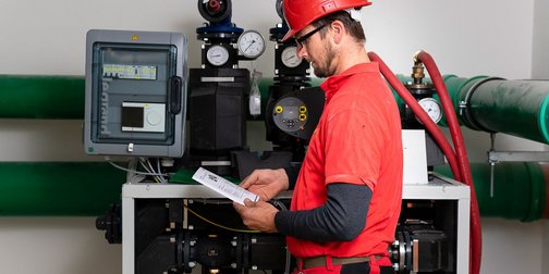 Ein Arbeiter in einem roten Hemd und Helm studiert eine Anleitung vor einer komplexen Maschinenanordnung mit Rohren.