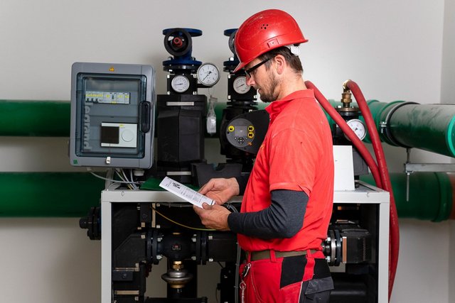 Ein Arbeiter in einem roten Hemd und Helm studiert eine Anleitung vor einer komplexen Maschinenanordnung mit Rohren.