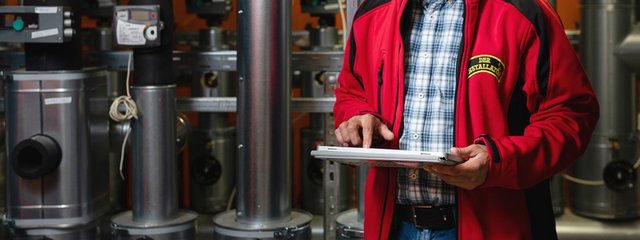 Der Installateur nutzt ein Tablet, um Daten zu überprüfen, umgeben von metallischen Rohren und Messgeräten in einer technischen Umgebung.