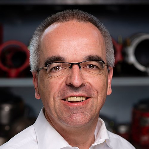 Der Mann mit grauen Haaren und Brille zeigt ein freundliches Lächeln, während er in einer industriellen Umgebung posiert.