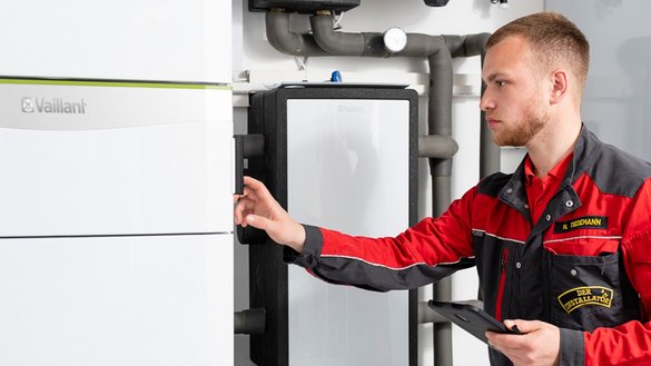 Ein junger Techniker in roter Arbeitskleidung überprüft ein Heizgerät und hält ein Tablet in der Hand.