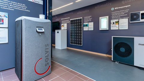 Ein moderner Ausstellungsraum mit einem grauen Wärmepumpengerät und einem Solarpanel an der Wand, umgeben von Informationsplakaten.