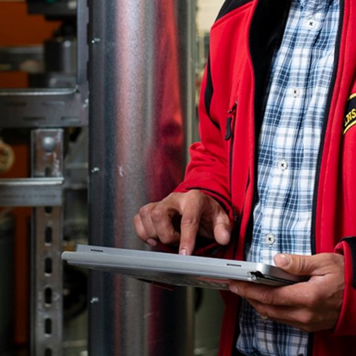 Ein Techniker in einer roten Jacke bedient ein Tablet, während er in einer industriellen Umgebung arbeitet.