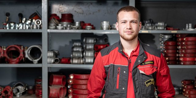 Ein junger Mann in einem roten Arbeitsanzug steht vor einem Regal voller technischer Ausrüstung und Werkzeuge.