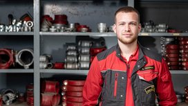 Ein junger Mann in einem roten Arbeitsanzug steht vor einem Regal voller technischer Ausrüstung und Werkzeuge.