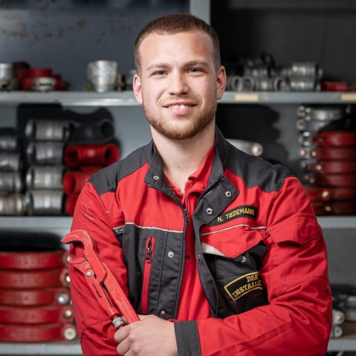 Ein junger Handwerker in roter Arbeitskleidung lächelt stolz, während er einen großen Schraubenschlüssel in der Hand hält.