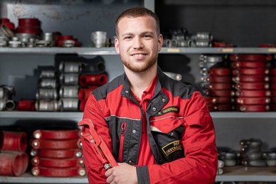 Ein junger Handwerker in roter Arbeitskleidung lächelt stolz, während er einen großen Schraubenschlüssel in der Hand hält.