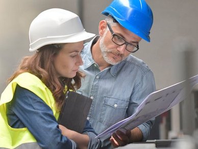 Ein Mann und eine Frau die einen Bauhelm tragen schauchen auf eine Technische Zeichnung