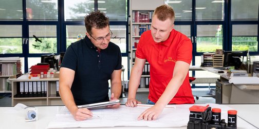 Zwei Männer stehen an einem Tisch und studieren einen Bauplan, während sie sich angeregt unterhalten.