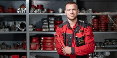 Ein junger Mechaniker in roter Arbeitskleidung lächelt stolz, während er einen Schraubenschlüssel in der Hand hält.