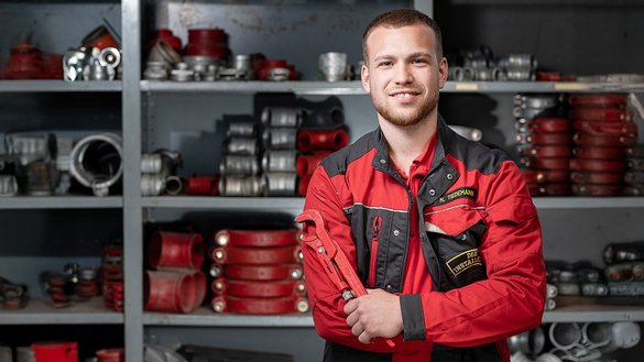 Ein junger Mechaniker in roter Arbeitskleidung lächelt stolz, während er einen Schraubenschlüssel in der Hand hält.