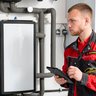 Ein junger Installateur in einem roten Arbeitsanzug überprüft eine Heizungsanlage und notiert Informationen auf einem Tablet.