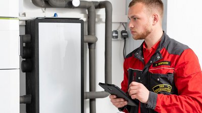 Ein junger Installateur in einem roten Arbeitsanzug überprüft eine Heizungsanlage und notiert Informationen auf einem Tablet.