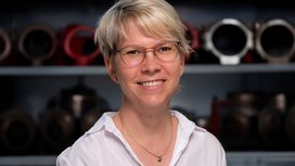 Die Frau mit kurzen, blonden Haaren und Brille strahlt Freude aus, während sie vor einer Reihe von Maschinen steht.