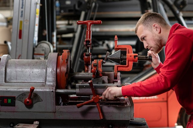 Ein junger Mann in einem roten Sweatshirt arbeitet konzentriert an einer Maschine in einer Werkstatt.