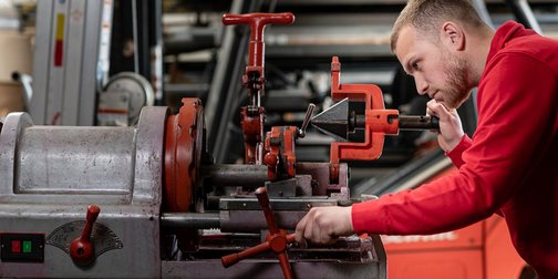 Ein junger Mann in einem roten Sweatshirt arbeitet konzentriert an einer Maschine in einer Werkstatt.