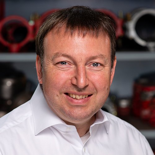 Ein lächelnder Mann mit kurzen, braunen Haaren trägt ein weißes Hemd und steht vor einer Werkstatt mit Maschinen im Hintergrund.