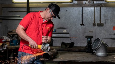Ein Handwerker in einem roten Shirt schneidet ein Metallrohr mit einer Winkelschleifmaschine, Funken fliegen auf.