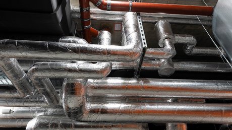 Ein komplexes Netzwerk von silbernen Rohren und Leitungen, die durch einen industriellen Raum verlaufen, mit einem schwarzen Behälter im Hintergrund.