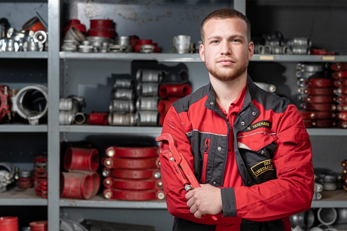 Ein junger Handwerker in roter Arbeitskleidung steht mit einem Schraubenschlüssel in der Hand vor einem Regal voller Werkzeuge.