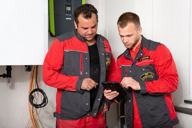 Zwei Handwerker in roten Arbeitsjacken betrachten gemeinsam ein Tablet in einem technischen Raum mit Kabeln im Hintergrund.