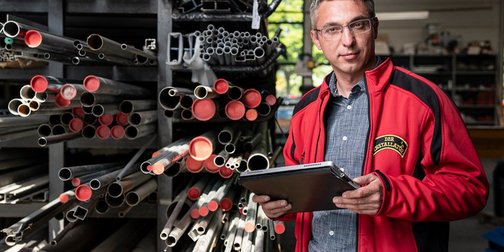 Ein Mann in einem roten Arbeitsjacke steht in einer Werkstatt, umgeben von verschiedenen Metallrohren und hält ein Tablet.
