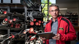 Fachmann in der Werkstatt mit Tablet und Rohren Ein Mann in einem roten Arbeitsjacke steht in einer Werkstatt, umgeben von verschiedenen Metallrohren und hält ein Tablet.