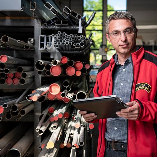 Ein Mann in einem roten Arbeitsjacke steht in einer Werkstatt, umgeben von verschiedenen Metallrohren und hält ein Tablet.