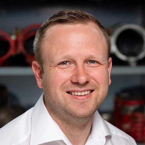 Ein lächelnder Mann mit kurzen, blonden Haaren trägt ein weißes Hemd und steht vor einer Werkstatt mit Maschinen im Hintergrund.