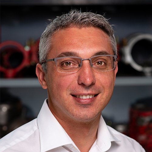 Ein lächelnder Mann mit Brille trägt ein weißes Hemd, während er vor einer Werkstatt mit Maschinen im Hintergrund steht.