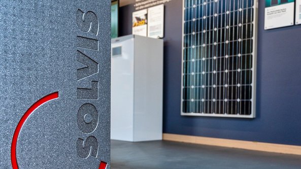 Ein graues Gerät mit dem Logo 'SOLVIS' steht im Vordergrund, während ein Solarpanel an der Wand sichtbar ist.
