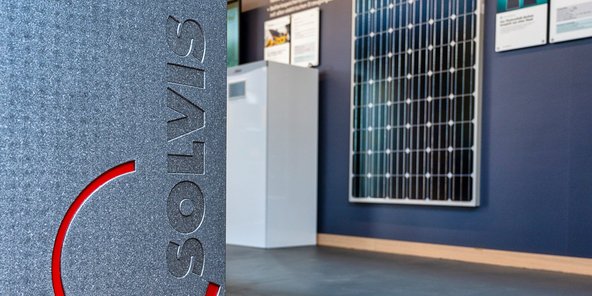 Ein graues Gerät mit dem Logo 'SOLVIS' steht im Vordergrund, während ein Solarpanel an der Wand sichtbar ist.