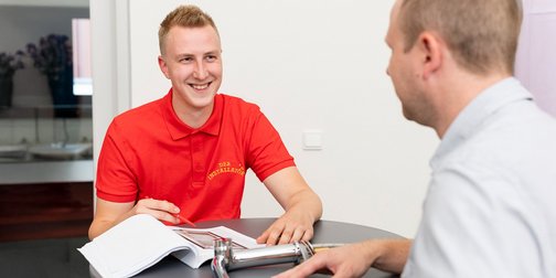 Ein junger Mann in einem roten Poloshirt lächelt, während er mit einem anderen Mann am Tisch spricht und Notizen macht.