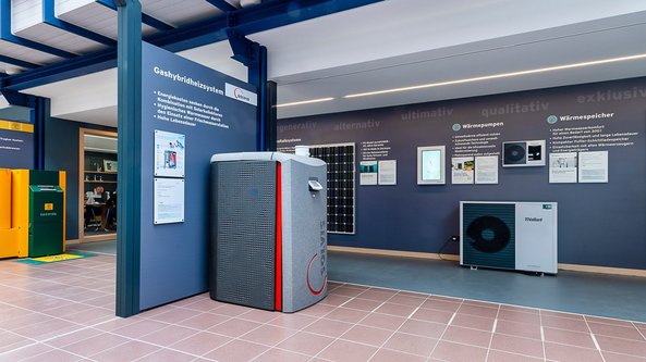 Ein moderner Ausstellungsraum zeigt verschiedene Heizsysteme, darunter einen grauen Wärmespeicher und eine Wärmepumpe.